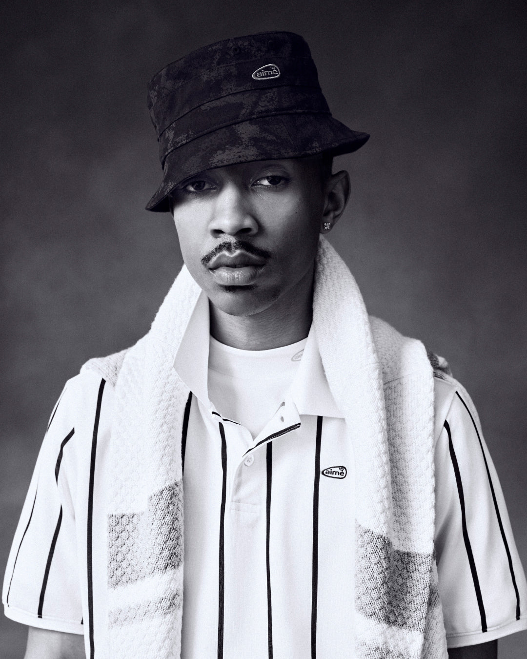 Black and white portrait of a man in an ALD Golf Bucket Hat, striped shirt, and towel over his shoulders.