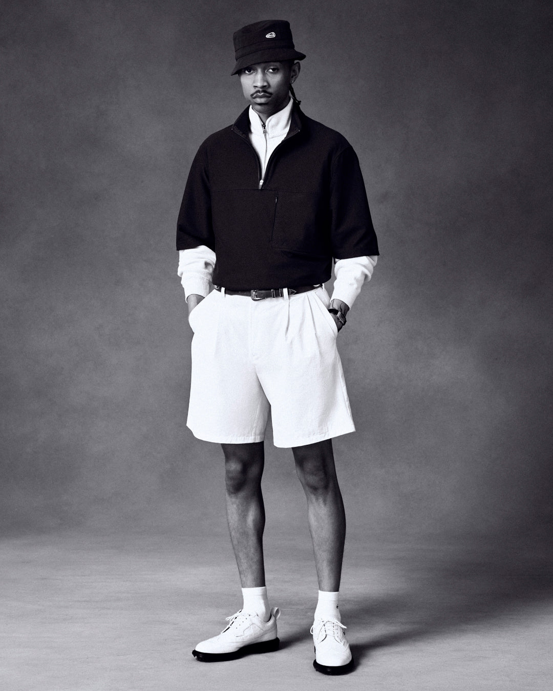 Man in ALD Golf Cordura Double Pleated Short, black bucket hat, dark pullover, long-sleeve shirt, and white shoes.