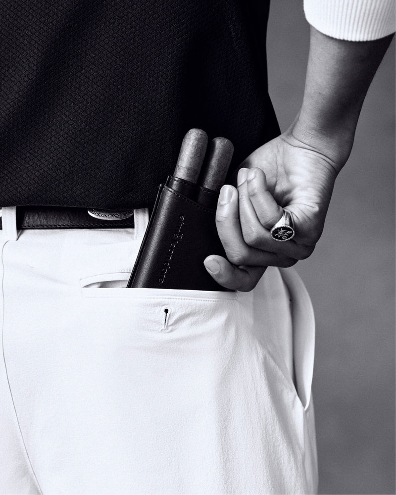 A person puts an ALD Golf Leather Cigar Case holding two cigars into the pocket of white pants, wearing a ring.