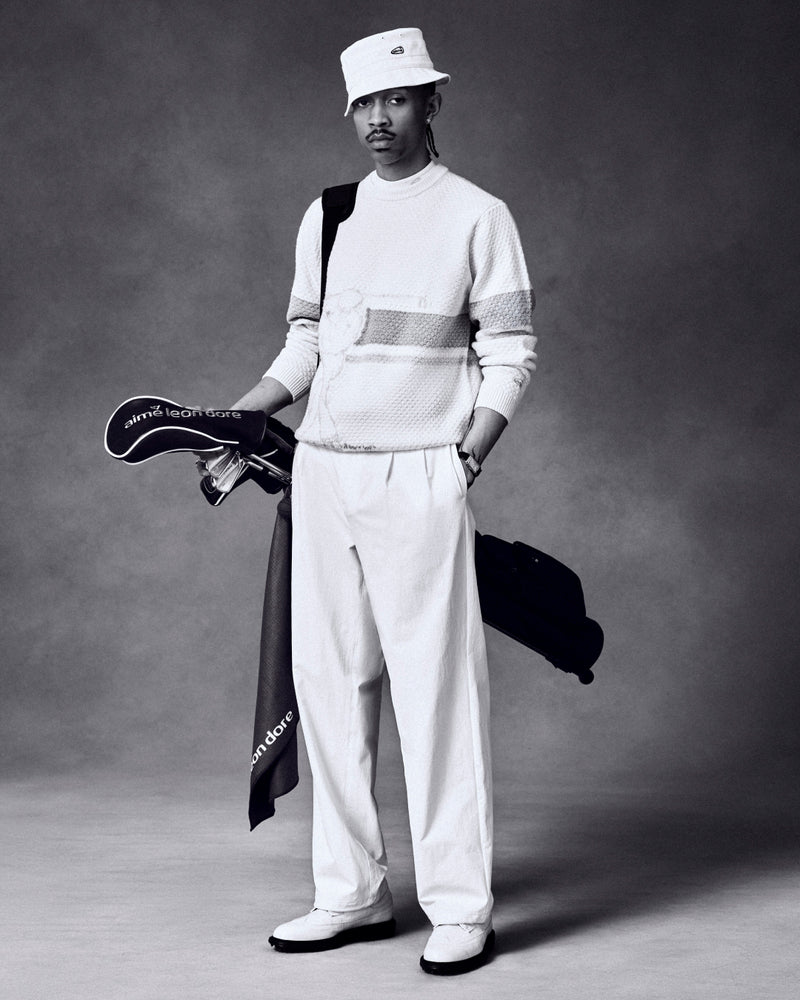 A man wears the ALD Golf Clubhouse Sweater, holding clubs and a bag, against a dark studio background.