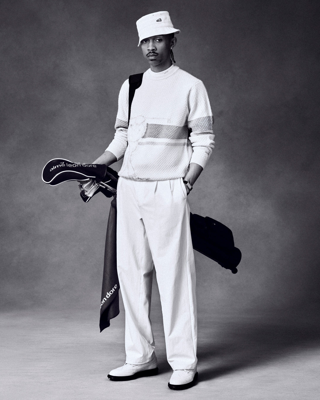 Man holding golf clubs with ALD Golf Driver Cover, dressed in light textured attire and bucket hat, against plain backdrop.