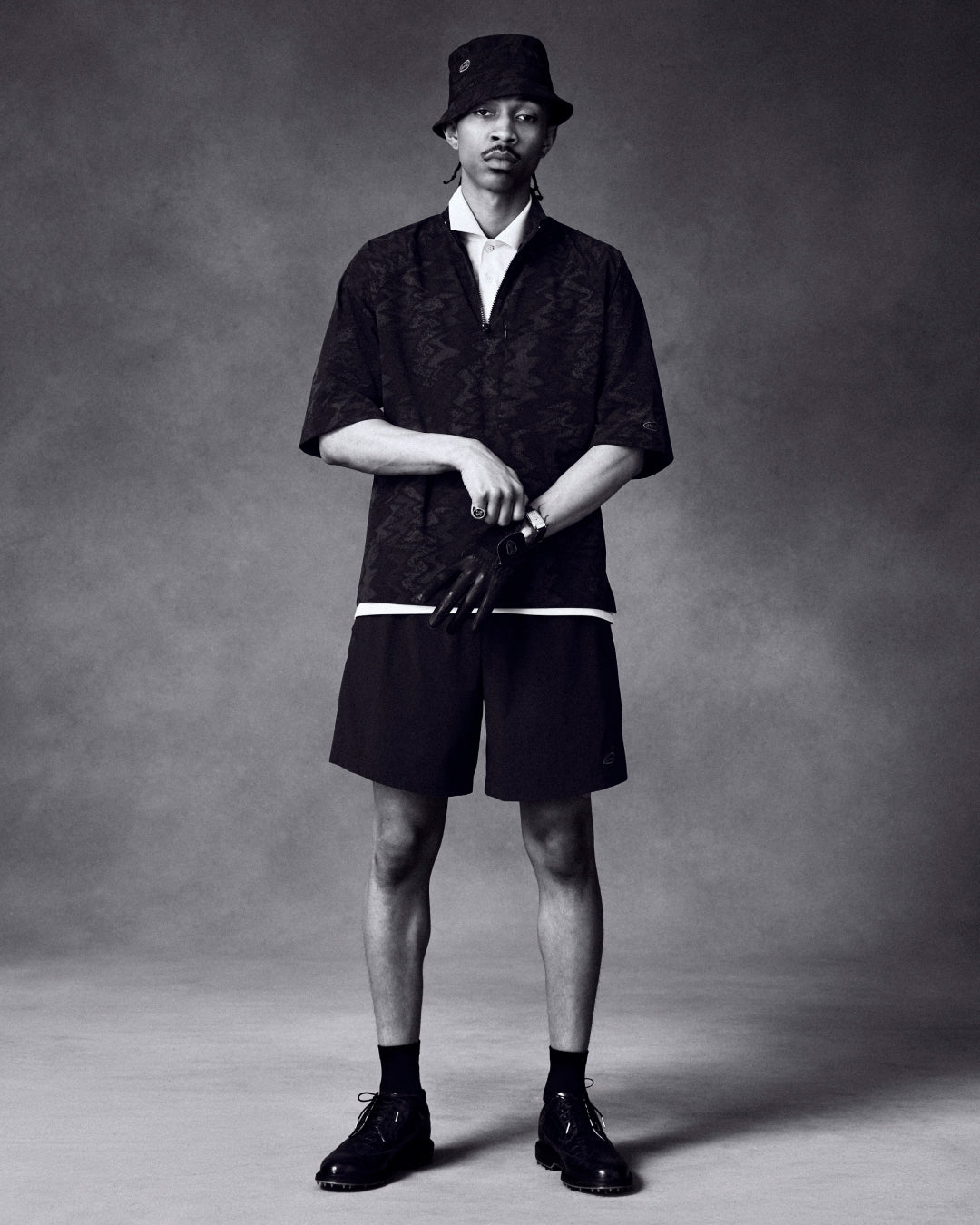 A man wears ALD Golf Performance Short with dark casual clothing, bucket hat, and gloves against a plain studio backdrop.