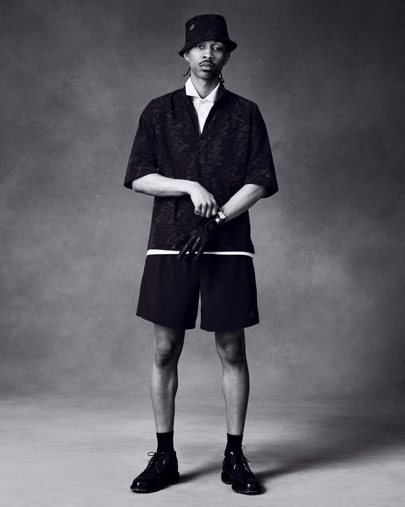 A man wears ALD Golf Performance Short with dark casual clothing, bucket hat, and gloves against a plain studio backdrop.