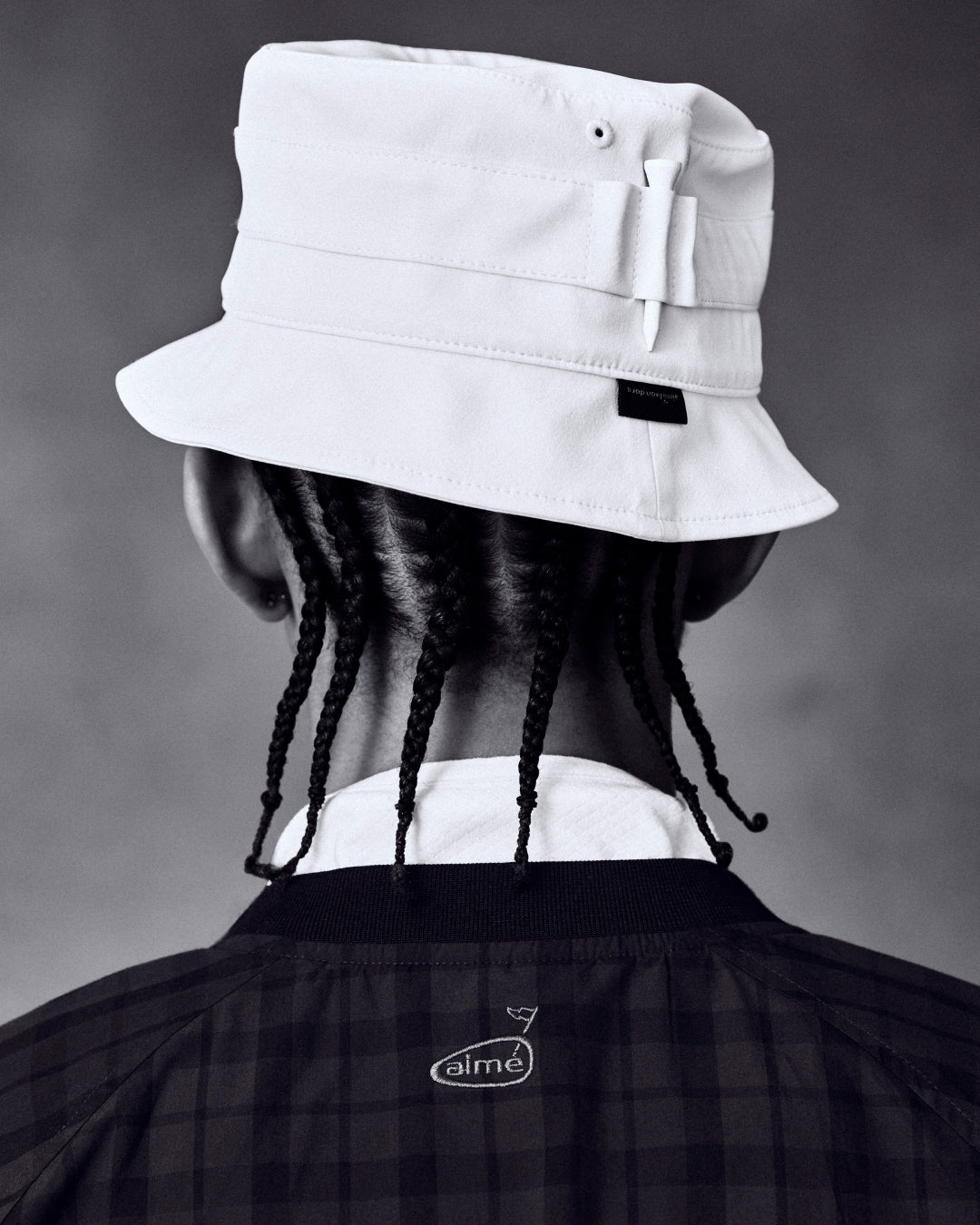 Person with braids wears the ALD Golf Bucket Hat and a dark checkered jacket, seen from behind against a neutral background.