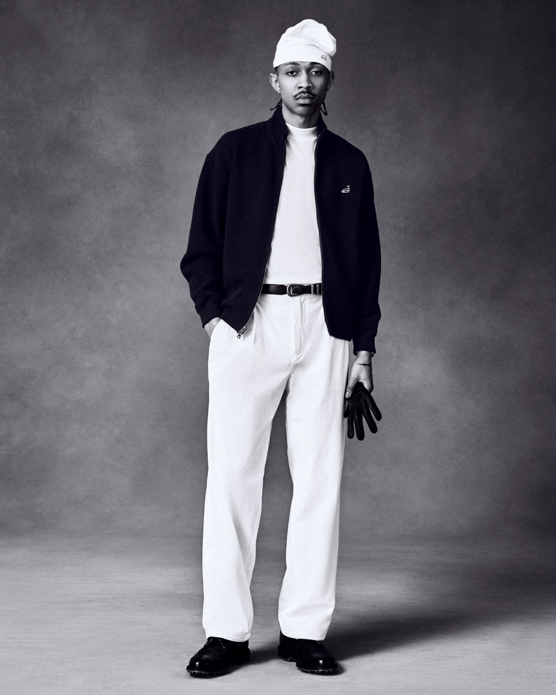 Man in a studio wears a white beanie, white pants, black jacket, and holds the ALD / FootJoy StaSof Heritage Glove.