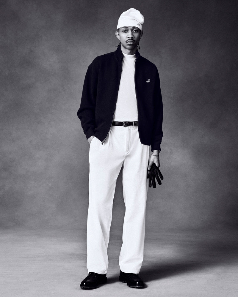 Man in a studio wears a white beanie, white pants, black jacket, and holds the ALD / FootJoy StaSof Heritage Glove.