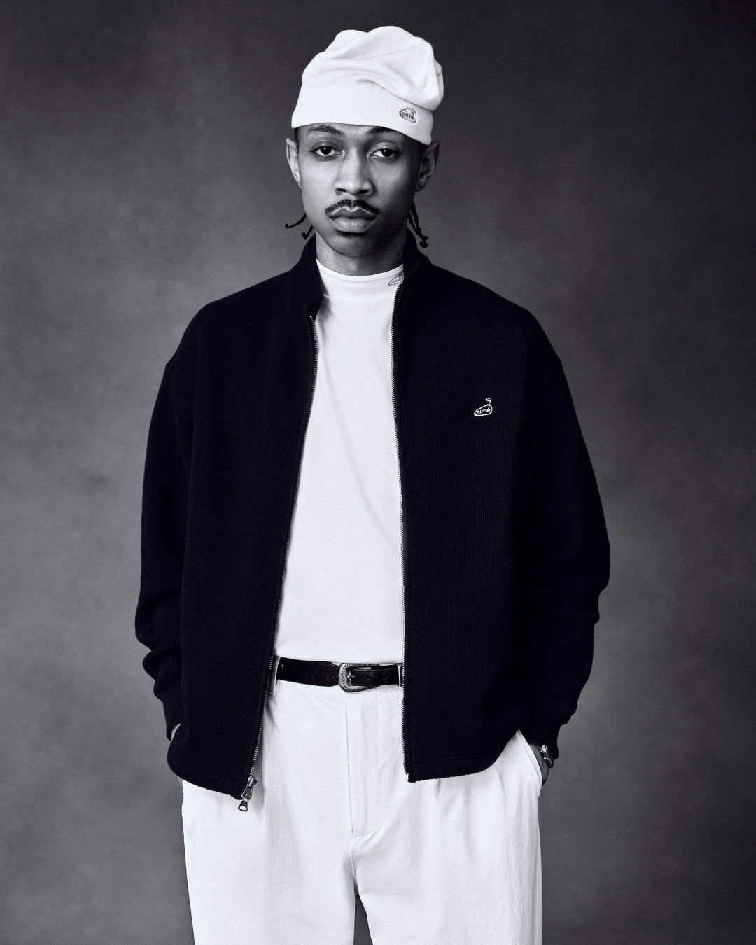Man in ALD Golf Performance Tee, white beanie, and black jacket stands with hands in pockets against a dark background.