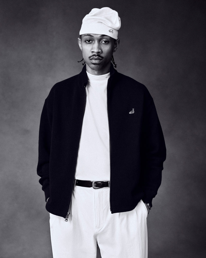Man in ALD Golf Performance Tee, white beanie, and black jacket stands with hands in pockets against a dark background.