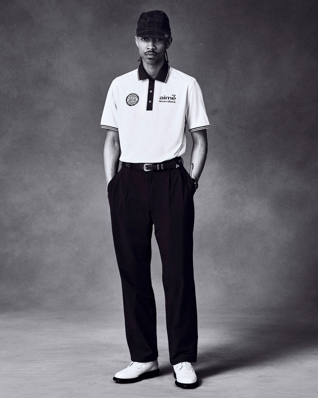 Man stands in studio, hands in pockets, wearing ALD Golf Cordura Double Pleated Pant, white polo, shoes, and a plaid cap.