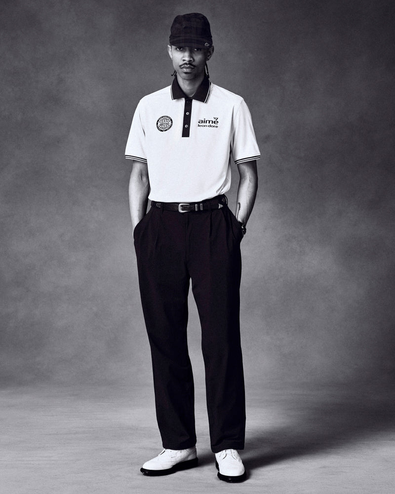 Man stands in studio, hands in pockets, wearing ALD Golf Cordura Double Pleated Pant, white polo, shoes, and a plaid cap.