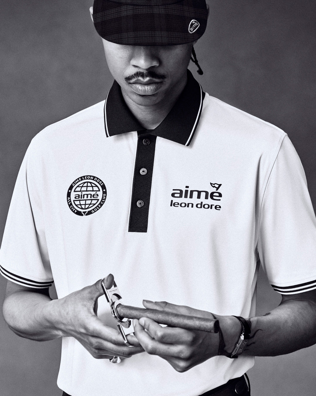Black and white photo of a man in an ALD Golf Tour Polo, holding a watch, wearing a plaid cap.