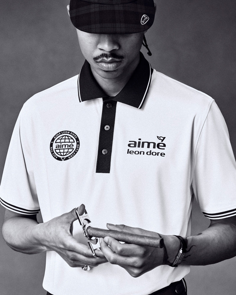 Black and white photo of a man in an ALD Golf Tour Polo, holding a watch, wearing a plaid cap.