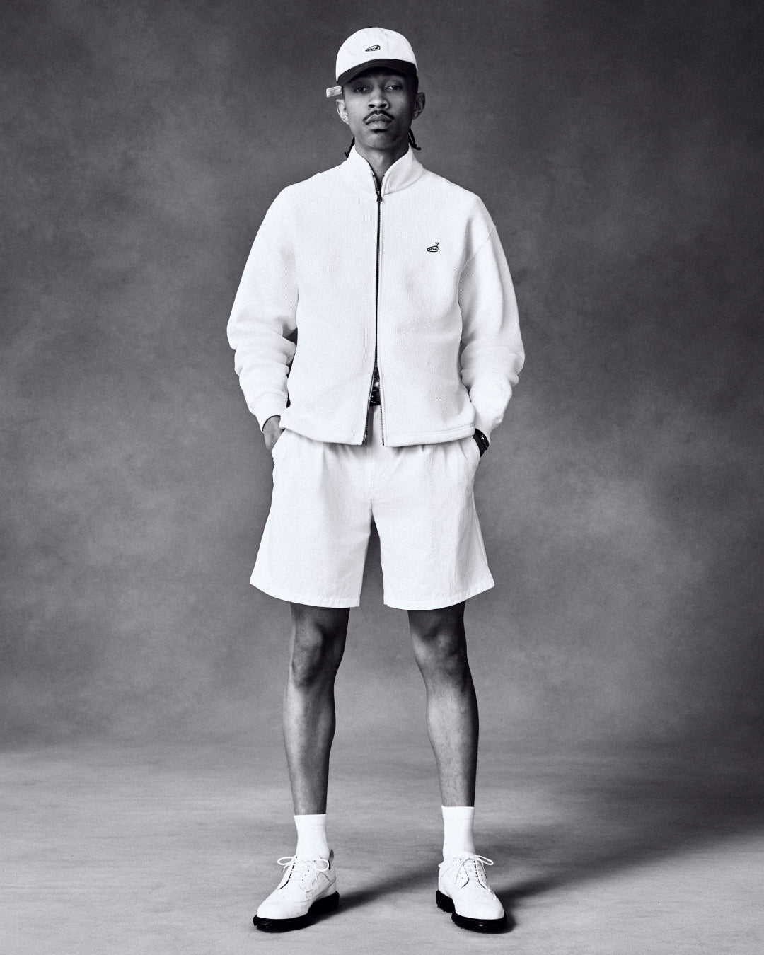 A man in the ALD Golf Piqué Full Zip Sweater stands hands-in-pockets against a neutral background.