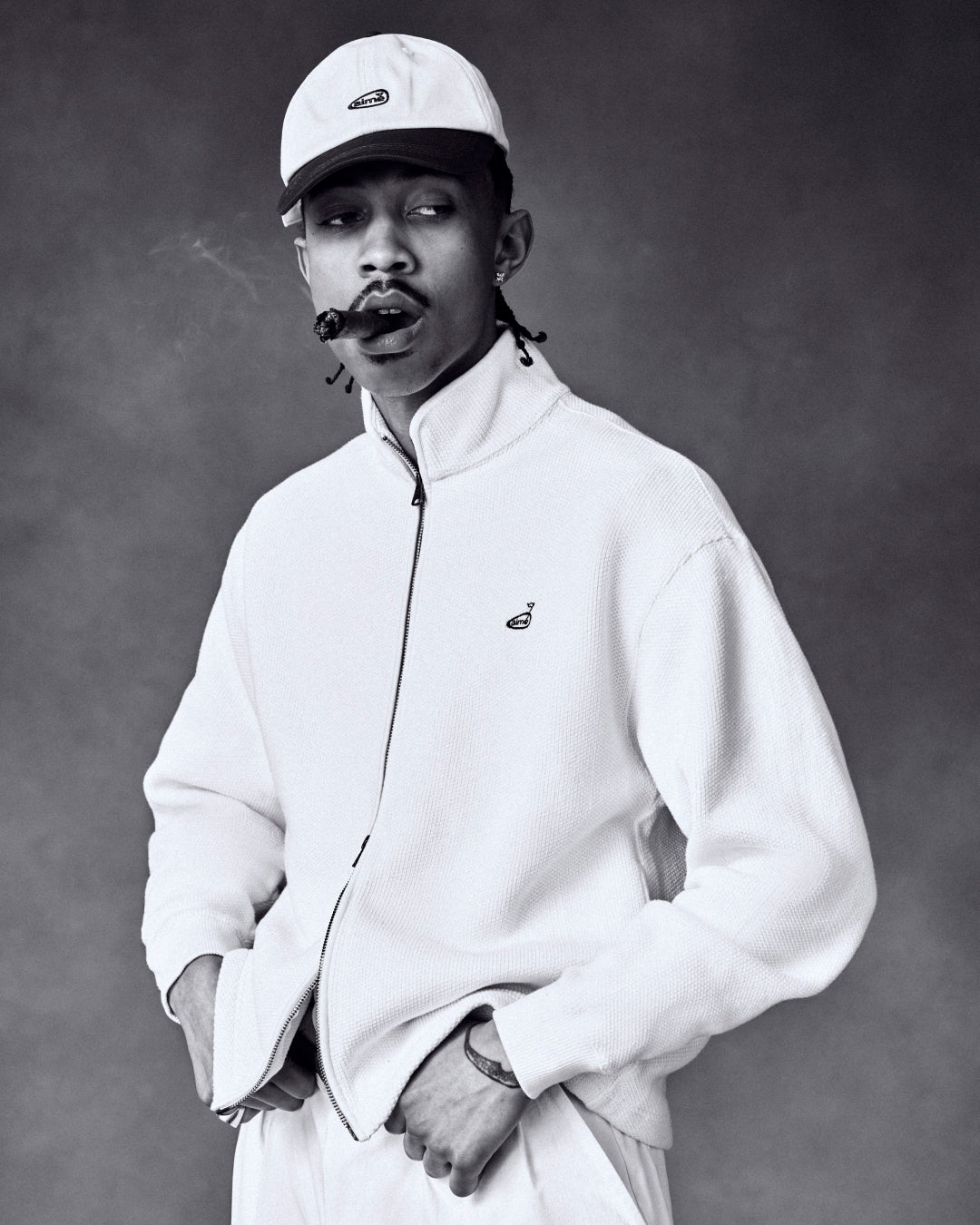 Black and white photo of a man in a light jacket and ALD Golf Logo Hat, smoking a cigar with his hands in his pockets.