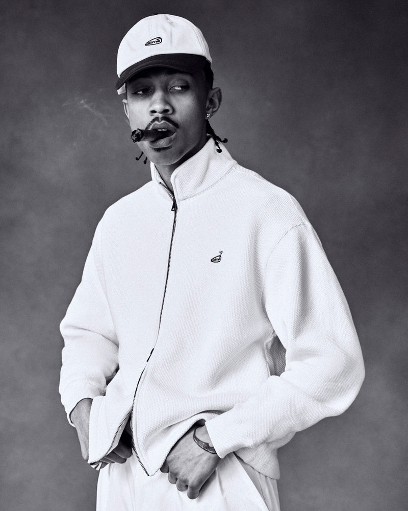 Black and white photo of a man in a light jacket and ALD Golf Logo Hat, smoking a cigar with his hands in his pockets.