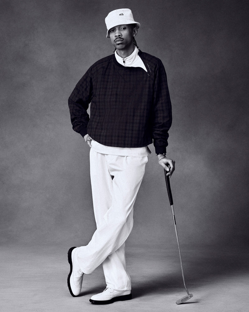 A man in an ALD Golf Blackwatch Pullover, bucket hat, and white pants holds a golf club against a dark studio backdrop.