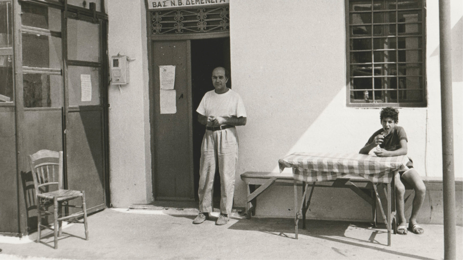 Two men standing outside a building with a table and chair in the foreground.