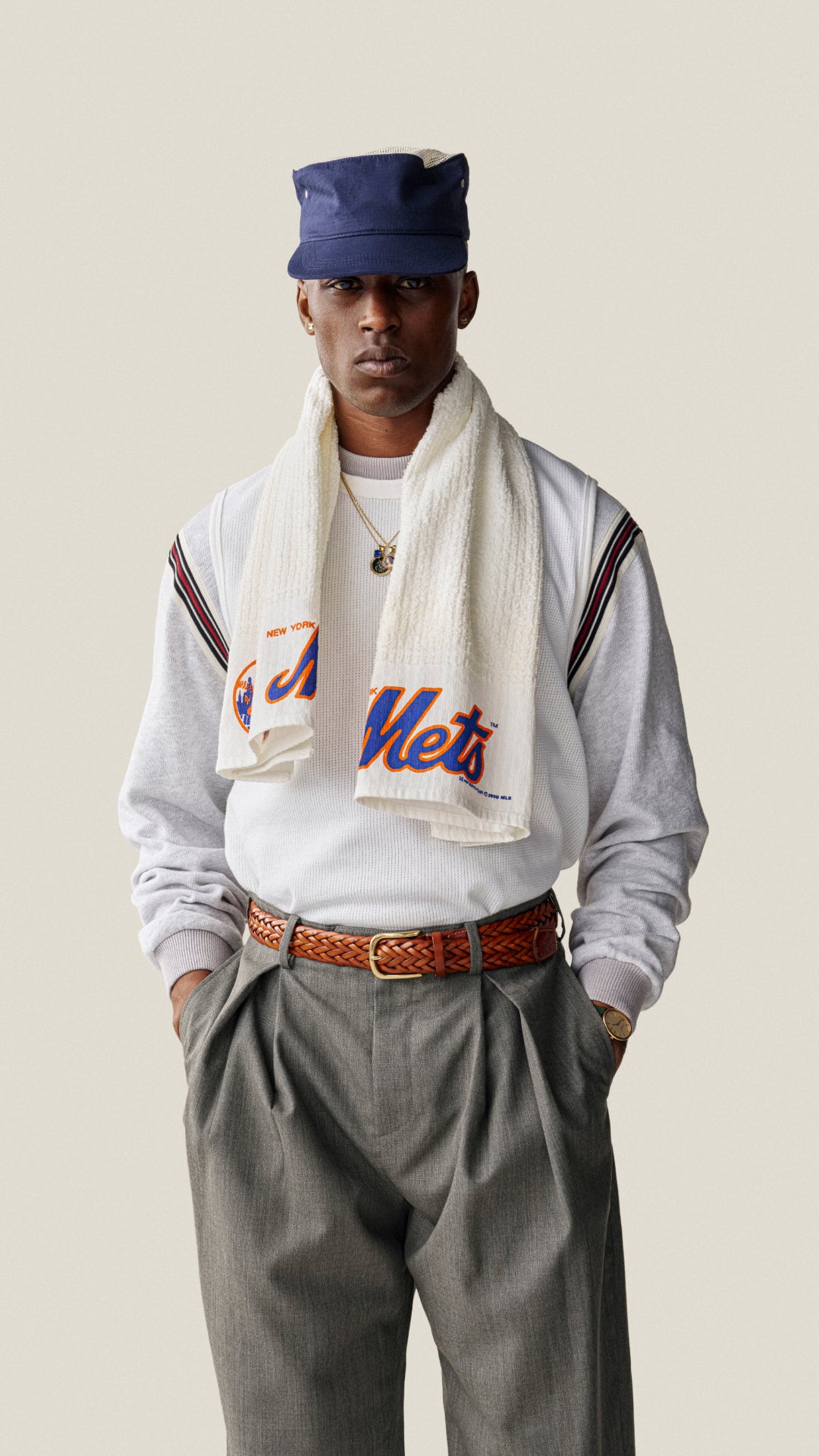 A man wearing a white sweatshirt with 'New York Mets' lettering, a white towel draped over his shoulders, a navy blue cap, and gray trousers, posed against a solid light beige background.