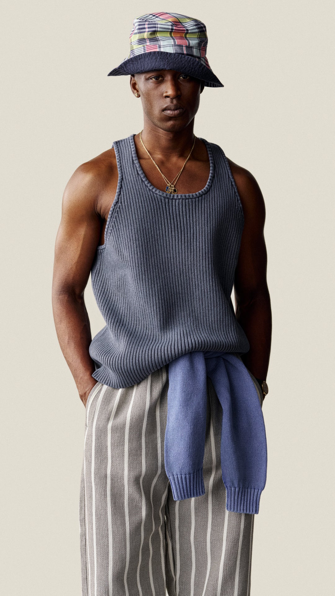 Man wearing a sleeveless blue top, plaid hat, and striped pants against a beige background