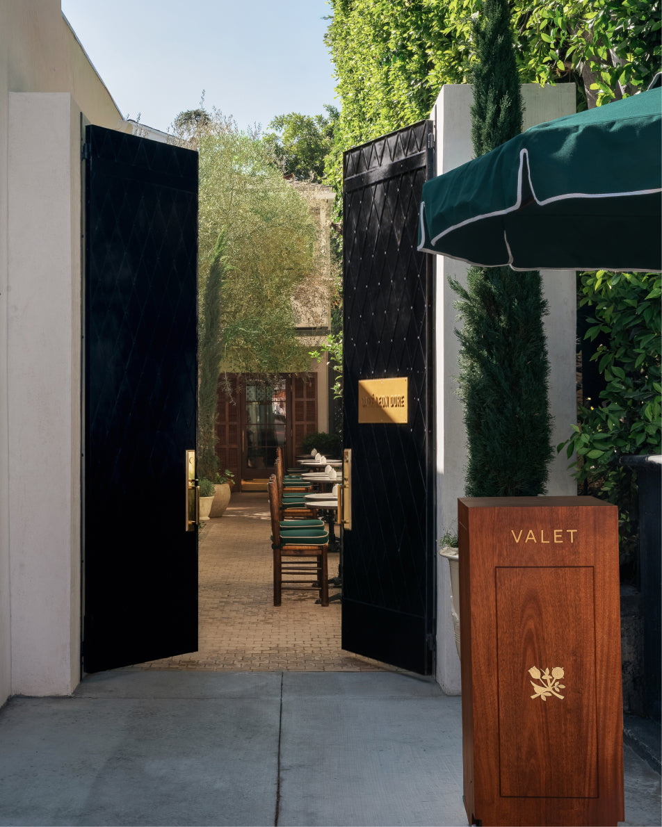 Open black doors leading to a valet area with chairs and tables, surrounded by greenery.