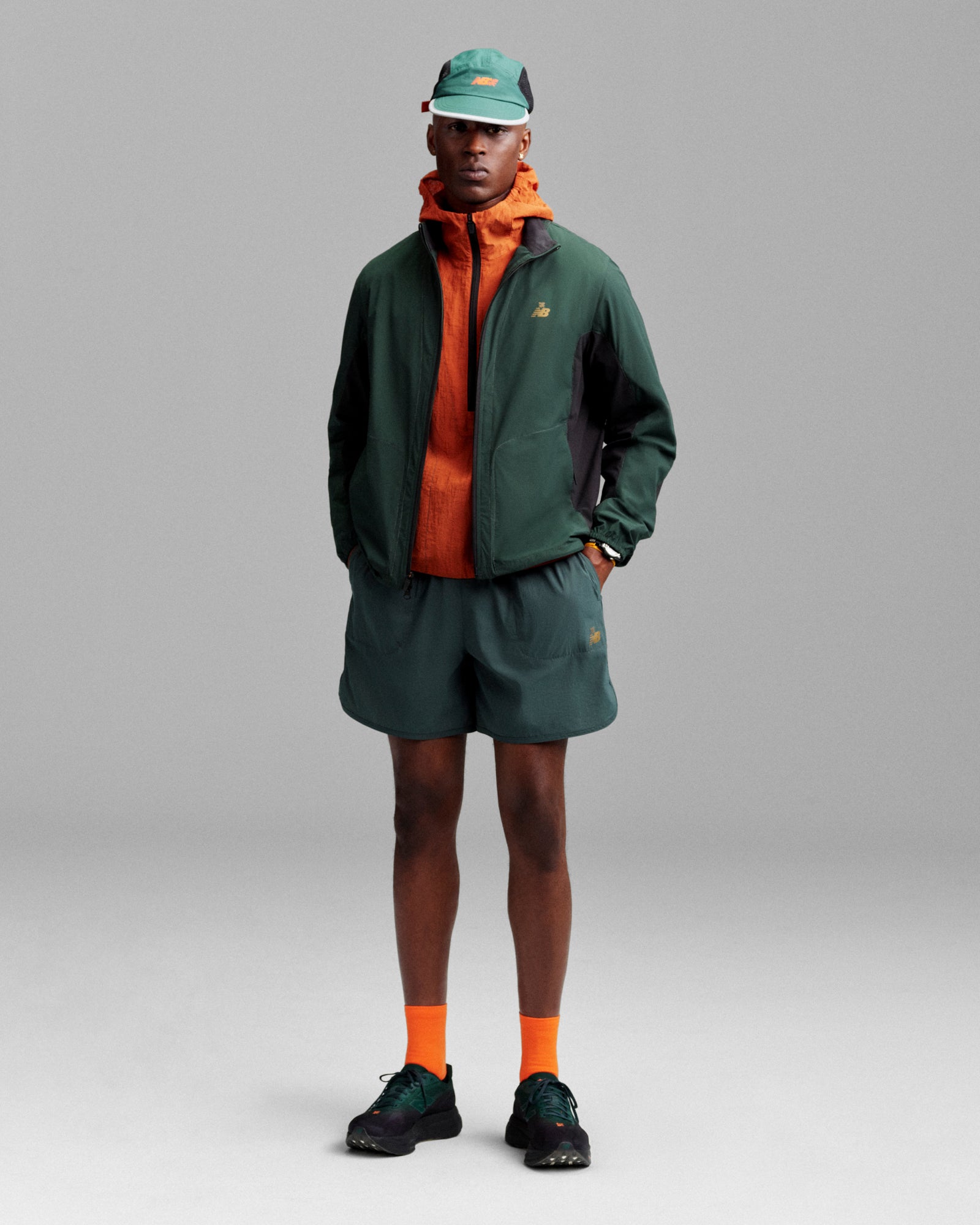A person wears ALD / New Balance Performance Lightweight Running Short with a green cap, orange jacket, and black sneakers.