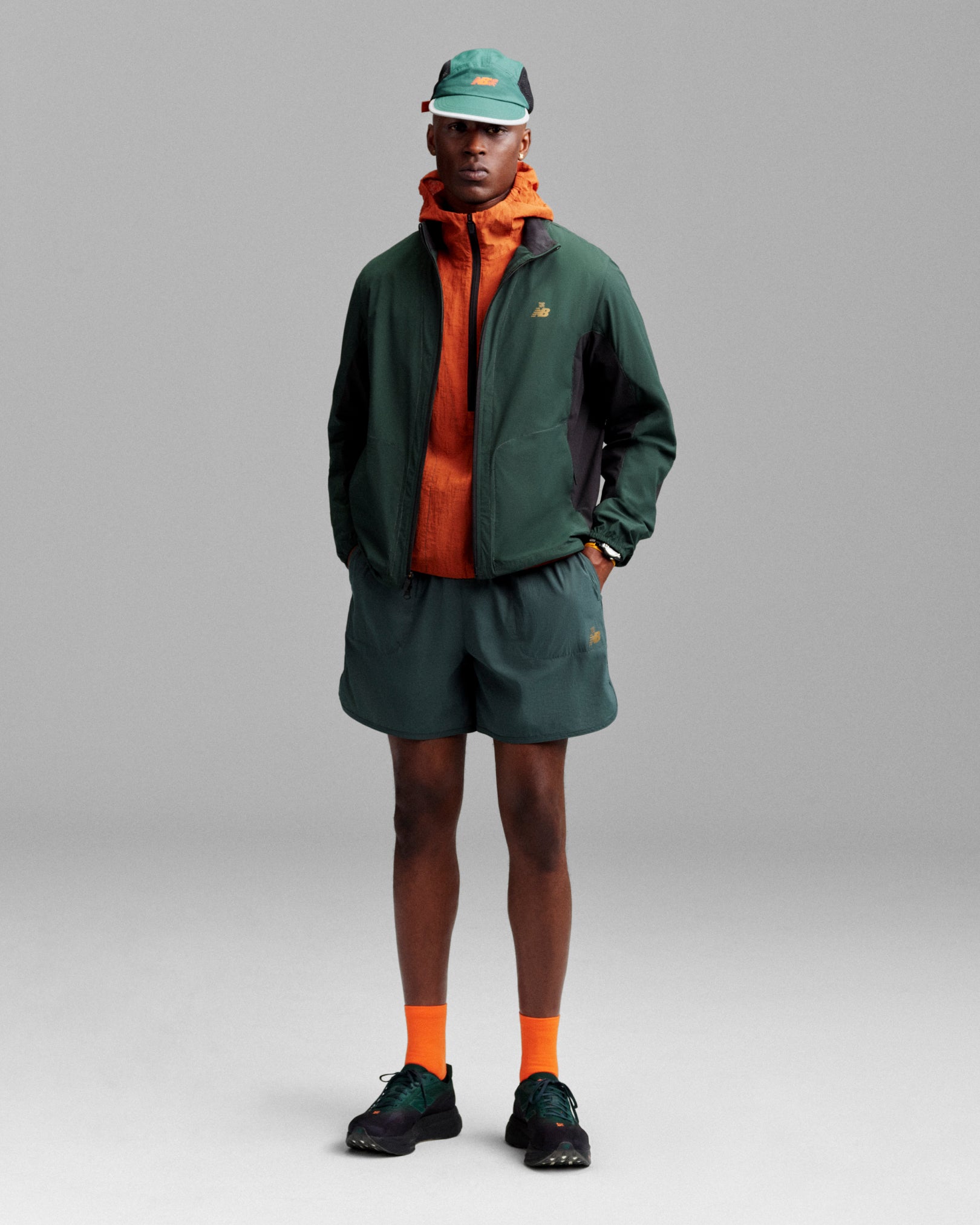 A person wears ALD / New Balance Performance Lightweight Running Short with a green cap, orange jacket, and black sneakers.