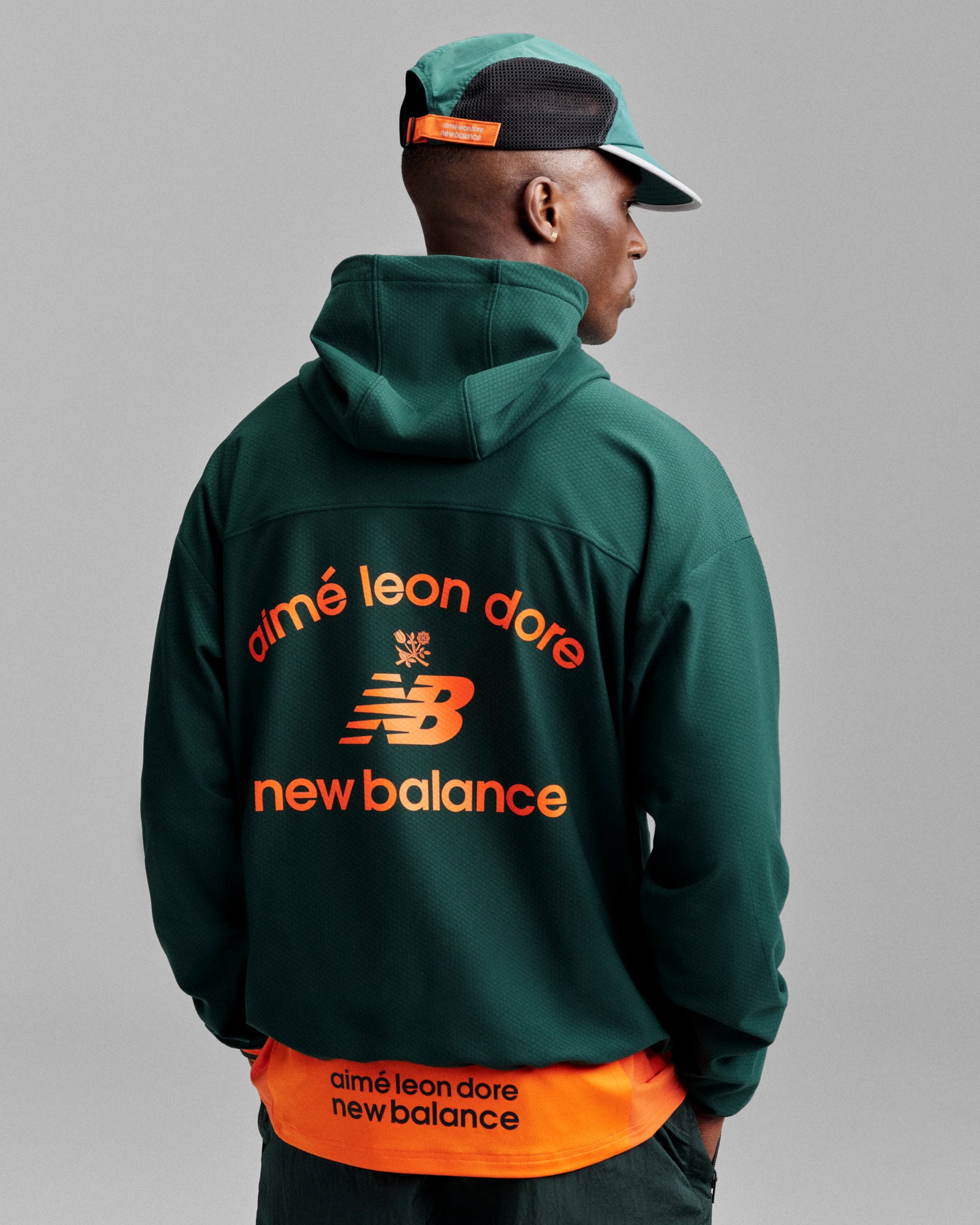 Man wearing an ALD / New Balance Full-Zip Warmup Hoodie and a green and orange cap, facing away.