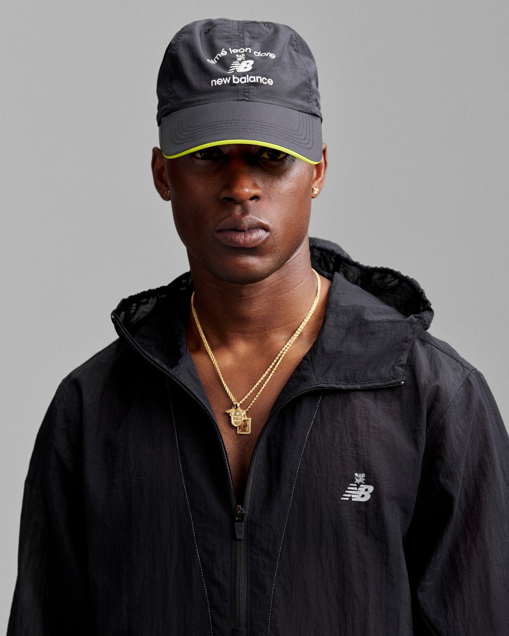 Man in an ALD / New Balance Running Hat and black jacket, wearing gold necklaces against a gray background.
