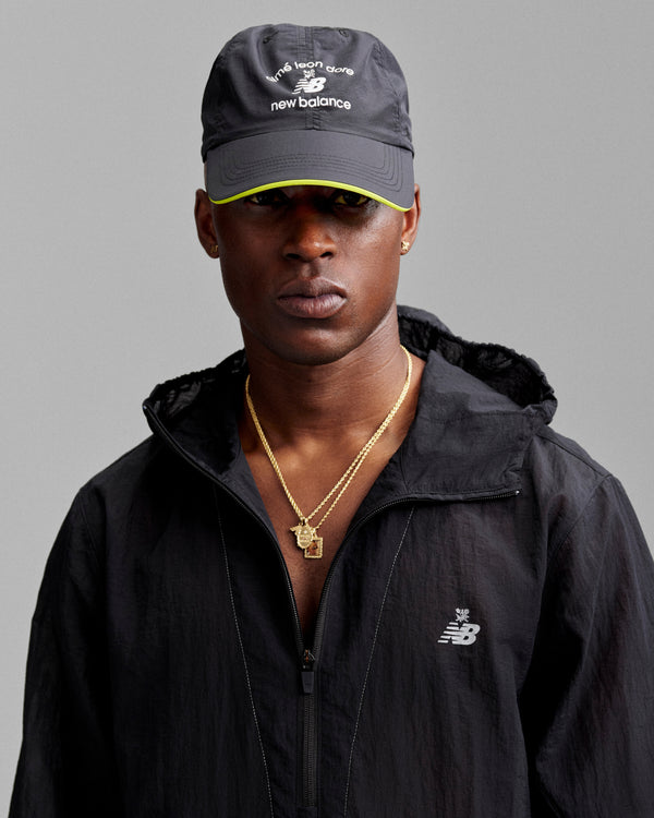 Man in an ALD / New Balance Running Hat and black jacket, wearing gold necklaces against a gray background.