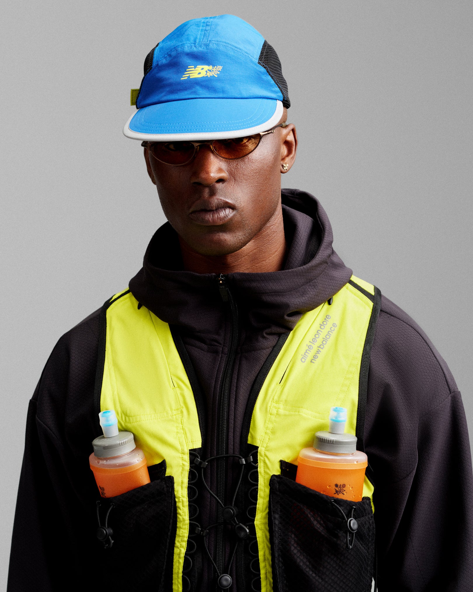 Person wearing an ALD / New Balance Mesh Sport Hat, yellow vest with bottles, dark hoodie, sunglasses, facing forward.