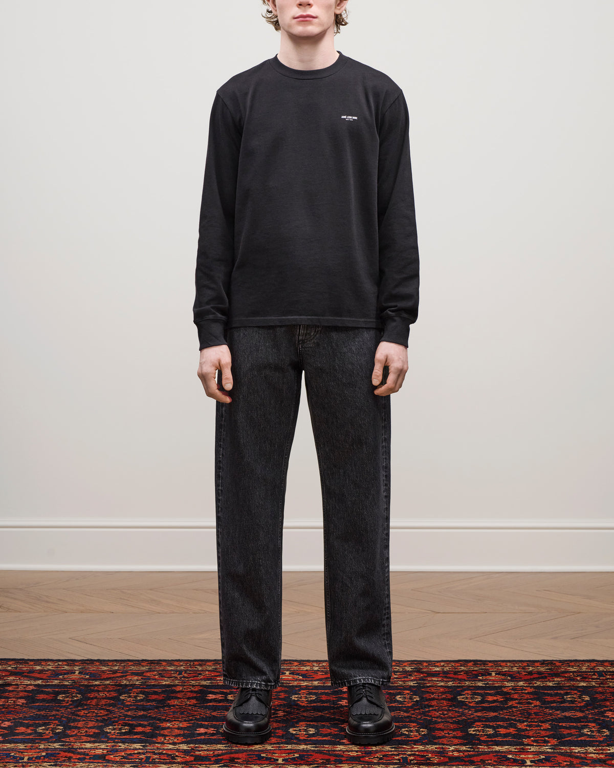 A person in a black Long-Sleeve Micro Logo Tee, dark jeans, and black shoes stands on a patterned rug indoors.