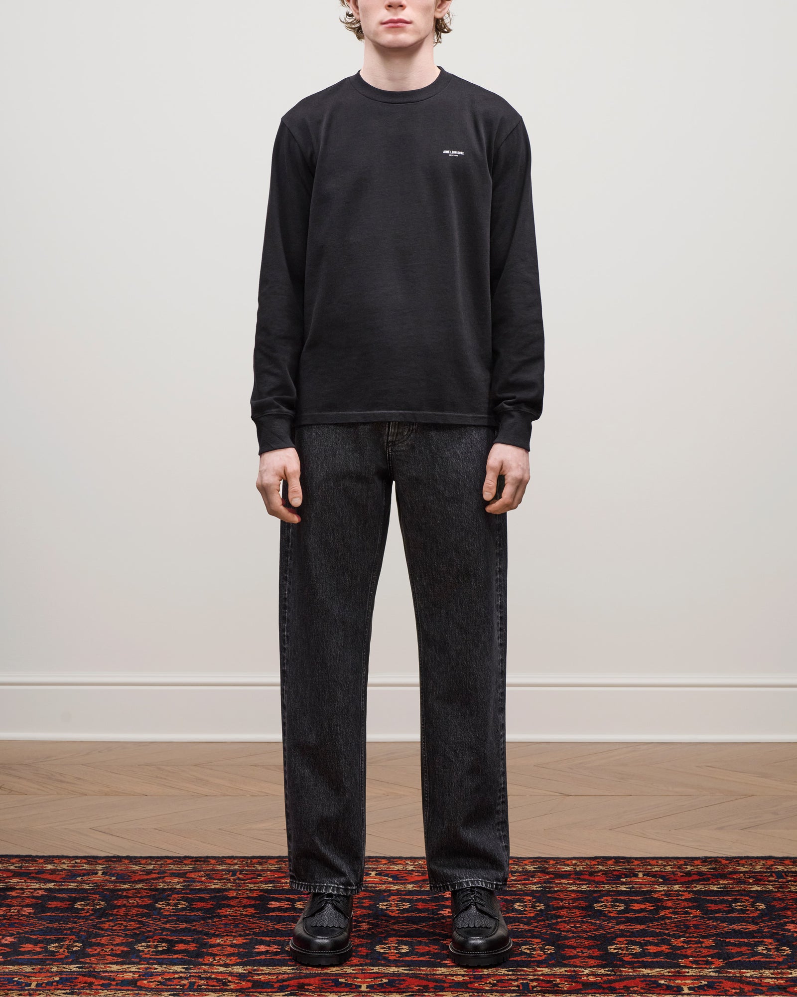 A person in a black Long-Sleeve Micro Logo Tee, dark jeans, and black shoes stands on a patterned rug indoors.