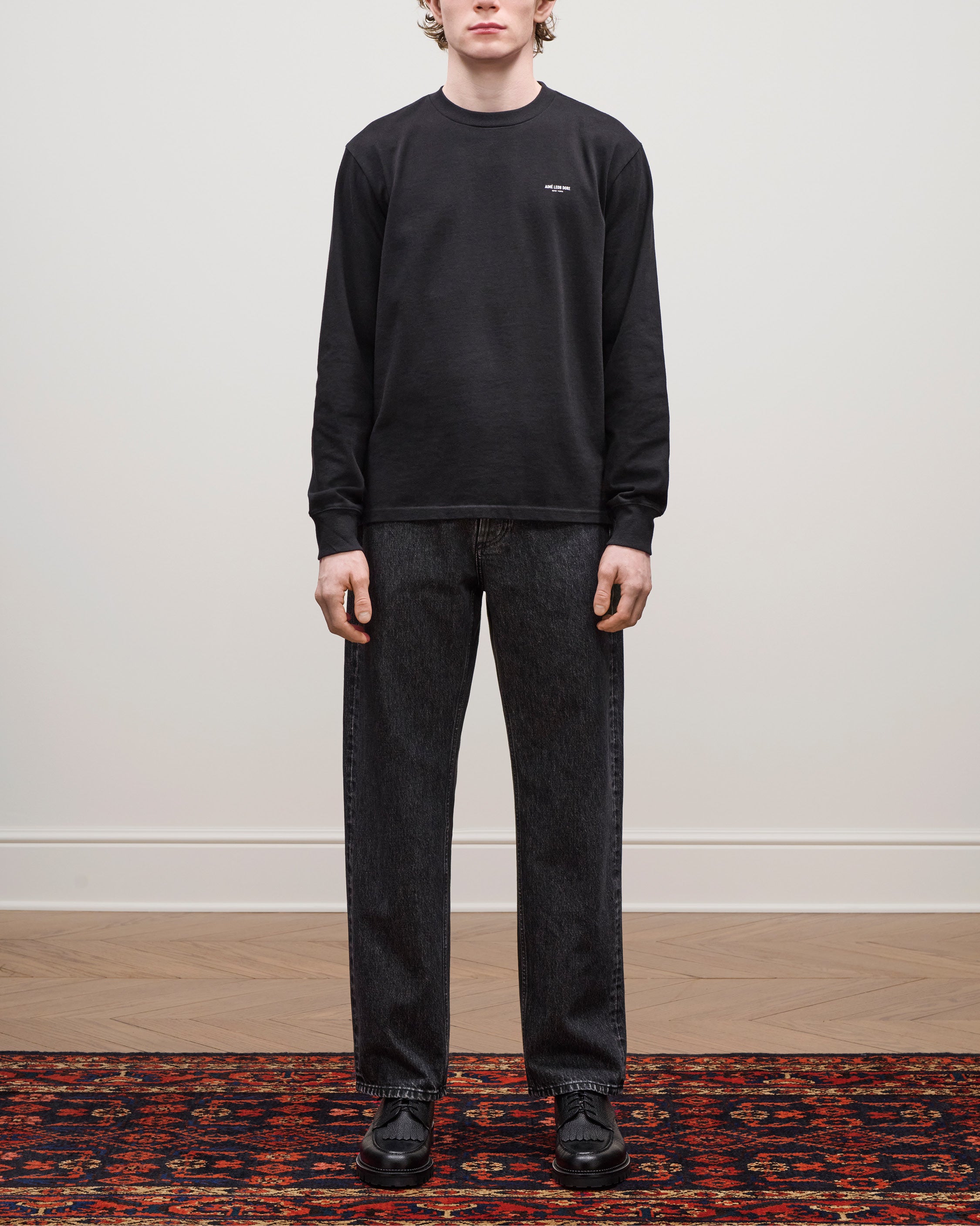 A person in a black Long-Sleeve Micro Logo Tee, dark jeans, and black shoes stands on a patterned rug indoors.