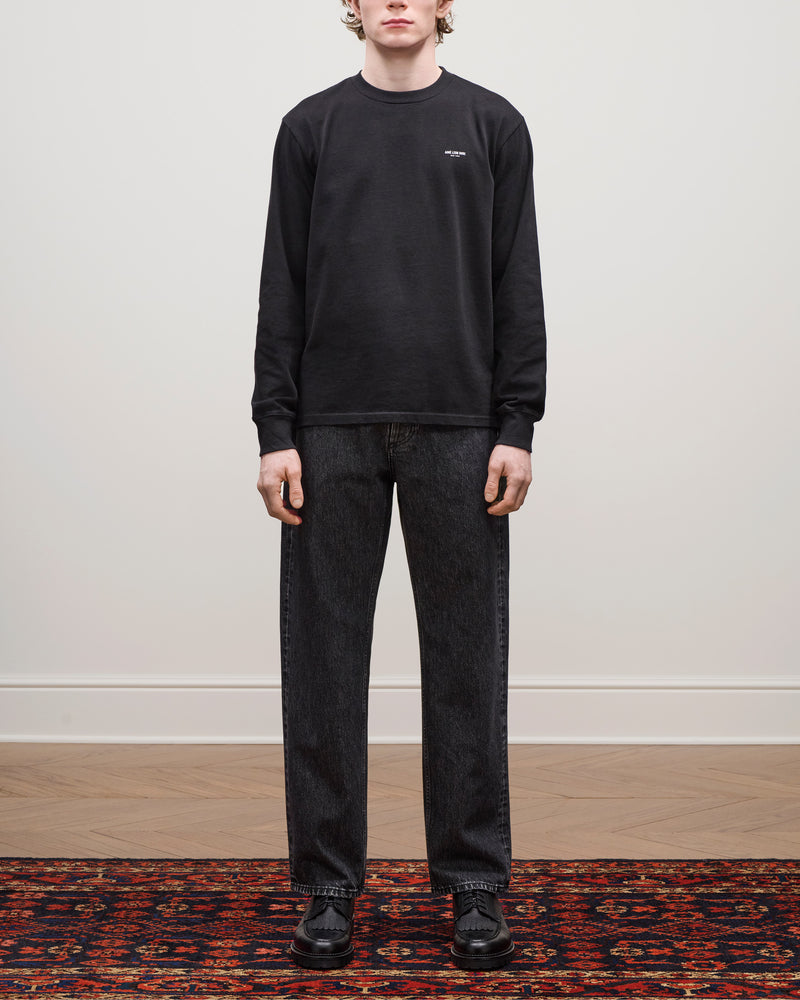 A person stands on a patterned rug wearing Relaxed Fit 5-Pocket Denim Pant, black sweatshirt, and black shoes.