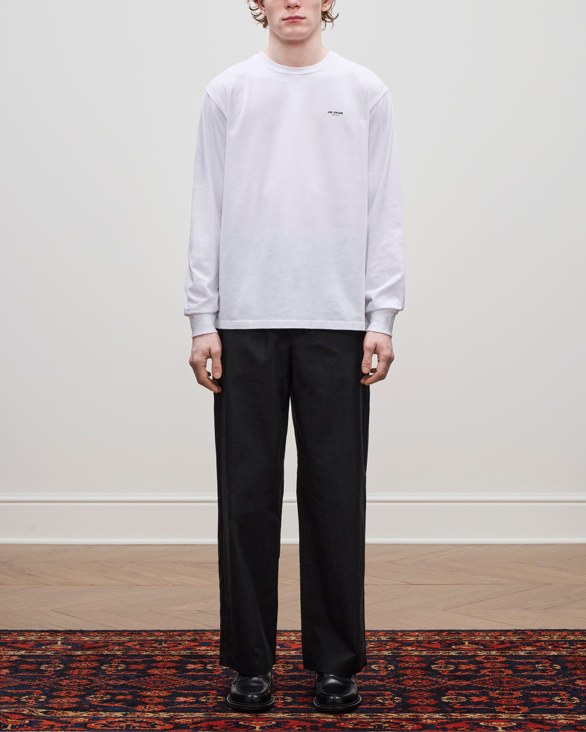 Person stands on a patterned rug wearing the Long-Sleeve Micro Logo Tee, black trousers, and black shoes.