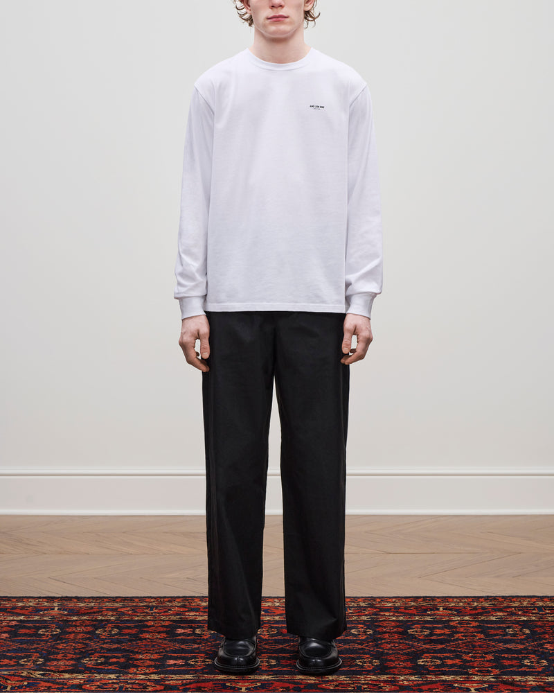 Person stands on a patterned rug wearing the Long-Sleeve Micro Logo Tee, black trousers, and black shoes.