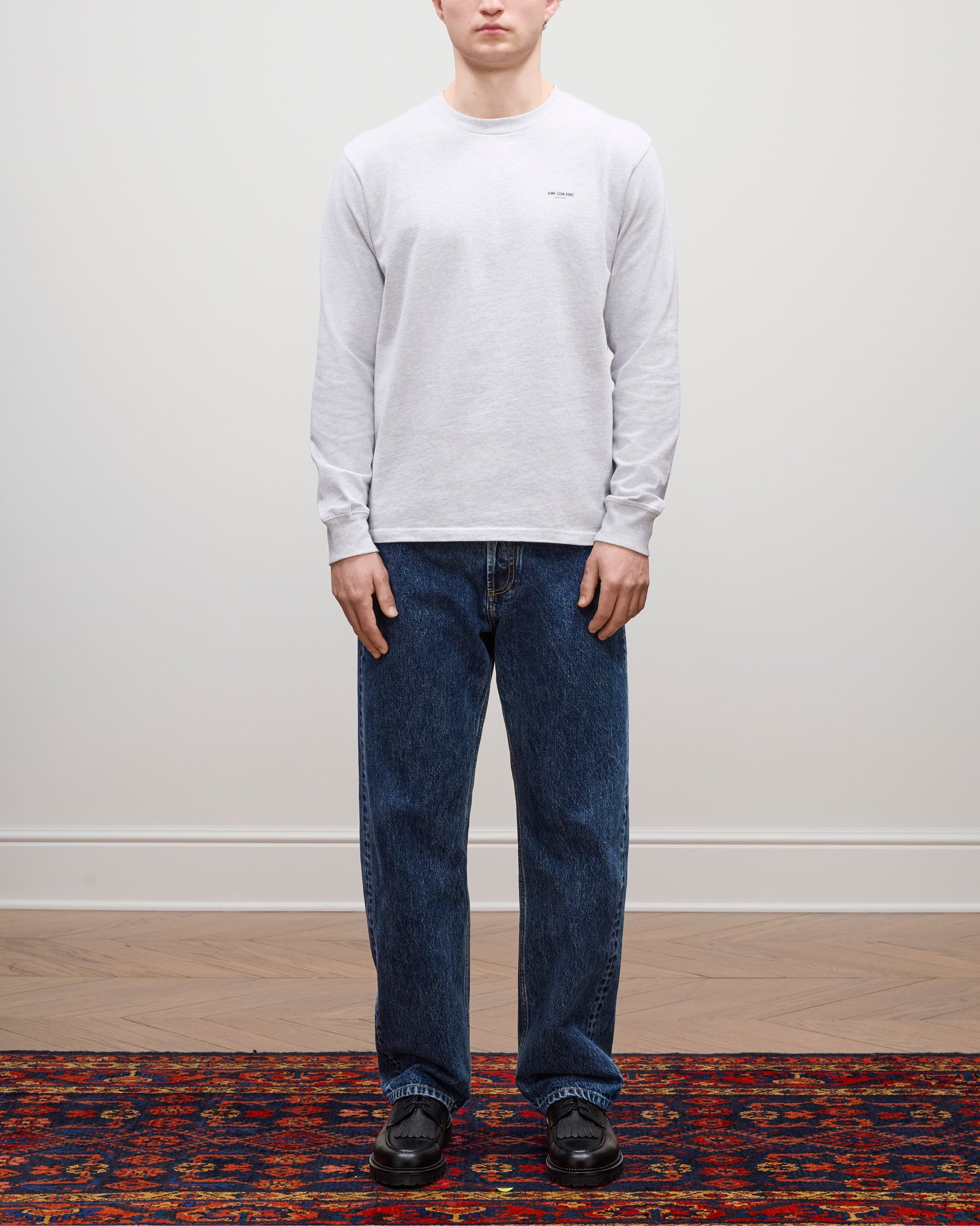 A person stands on a patterned rug wearing the Long-Sleeve Micro Logo Tee, blue jeans, and black shoes.