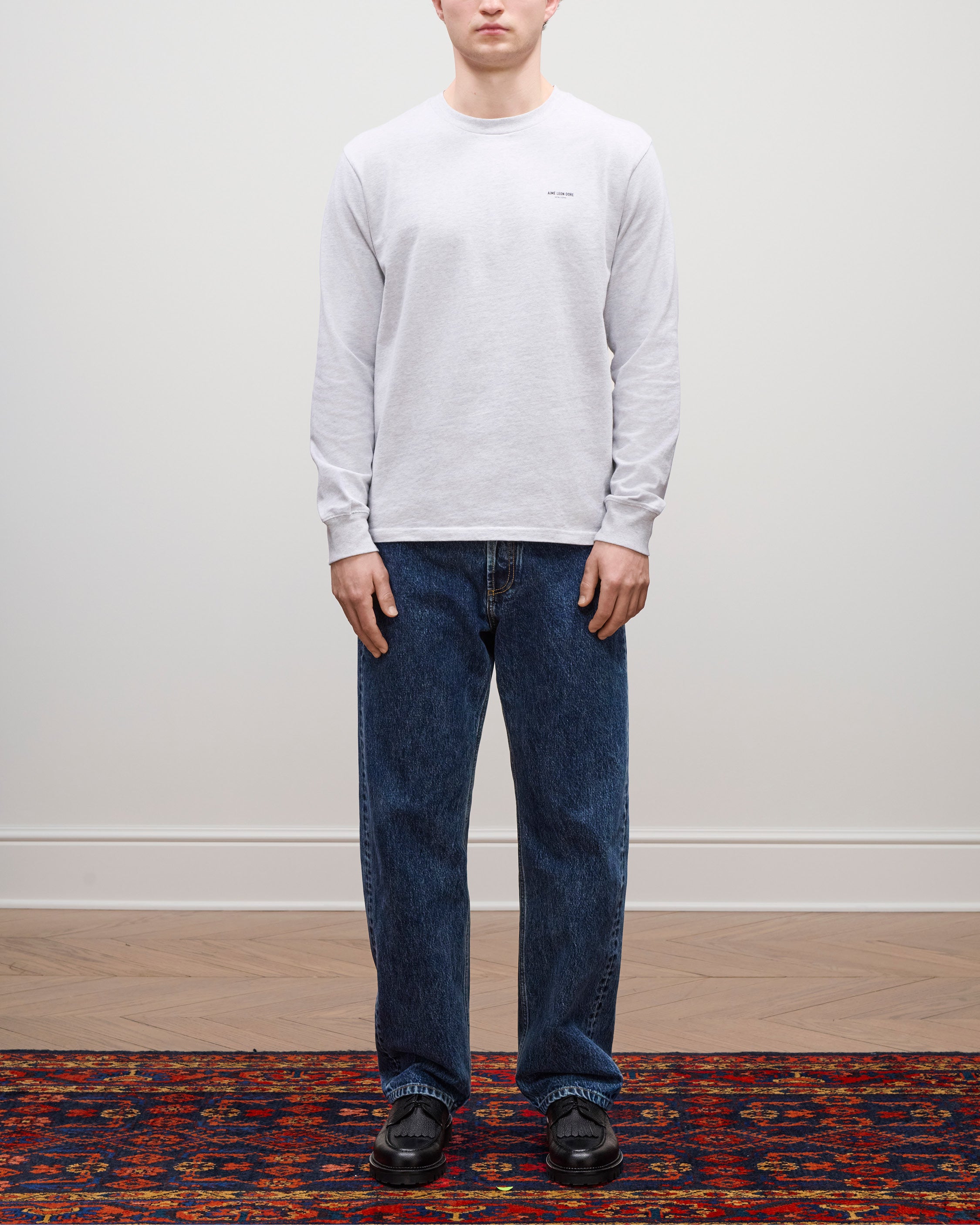 A person stands on a patterned rug wearing the Long-Sleeve Micro Logo Tee, blue jeans, and black shoes.