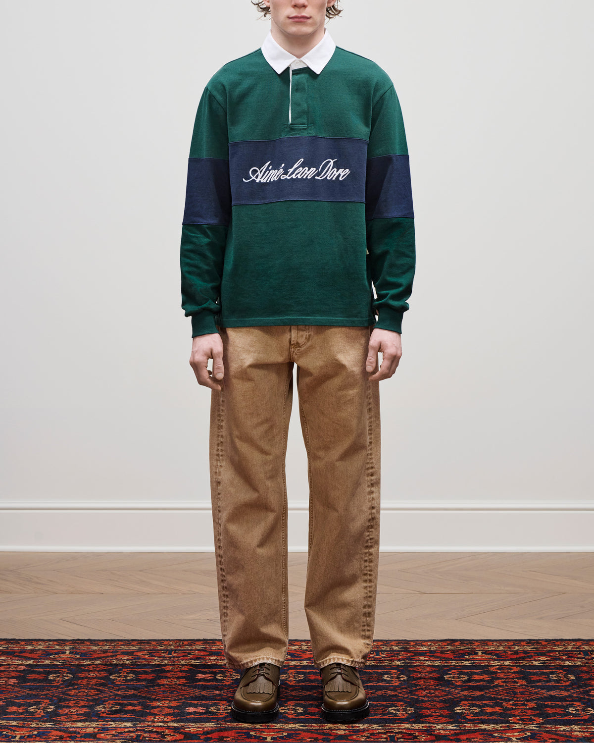Person wears a green Script Paneled Rugby shirt, tan pants, and brown shoes while standing on a rug.