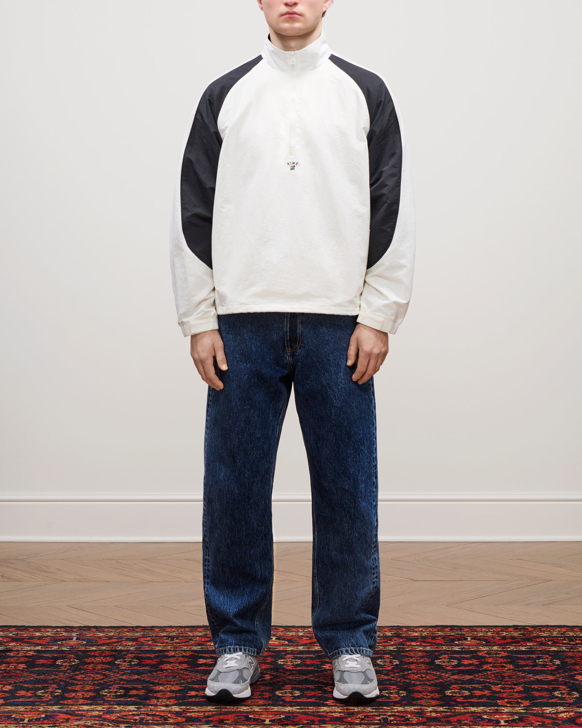 Person in a Crest Nylon Half-Zip Pullover, blue jeans, and gray sneakers standing on a patterned rug.