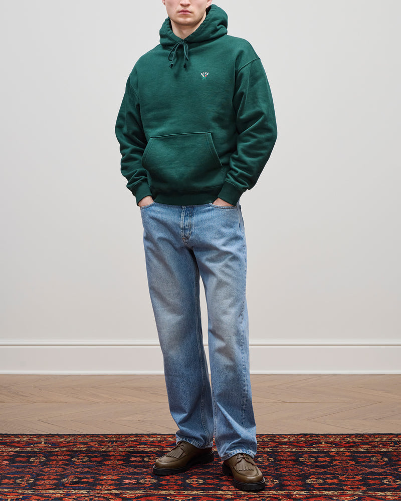 Person wears Relaxed Fit 5-Pocket Denim Pant, green hoodie, and brown shoes on a patterned rug by a plain wall.