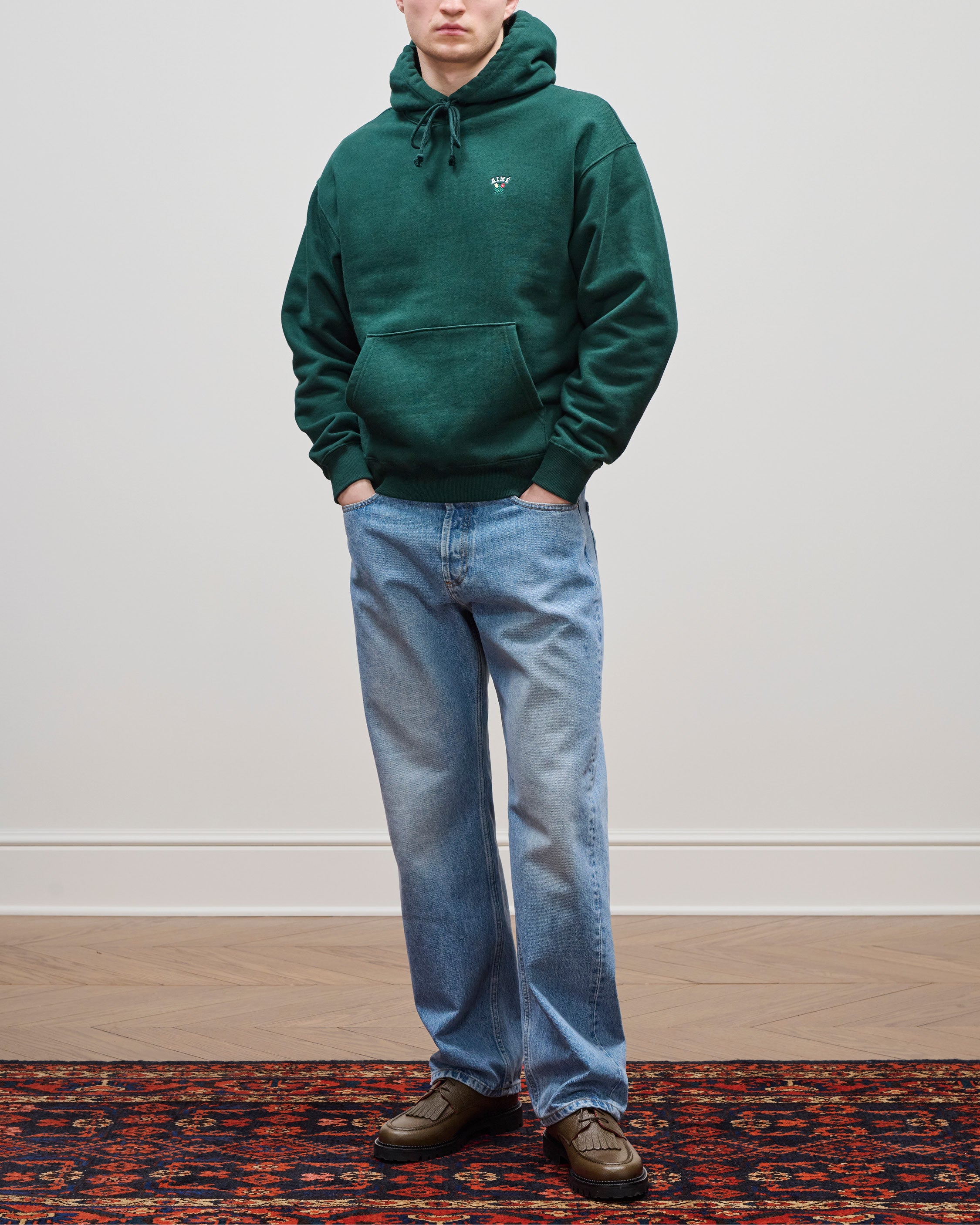 Person in a Crest Hoodie, light blue jeans, and brown shoes stands on a patterned red rug against a plain wall.
