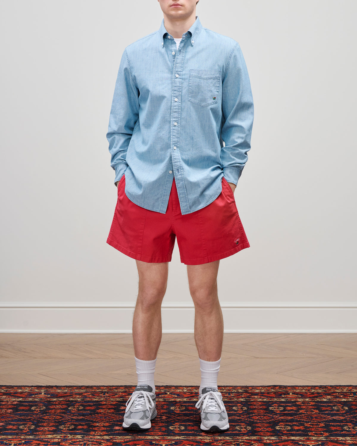 Someone wears a Crest Chambray Shirt, red shorts, white socks, and gray sneakers while standing on a rug.
