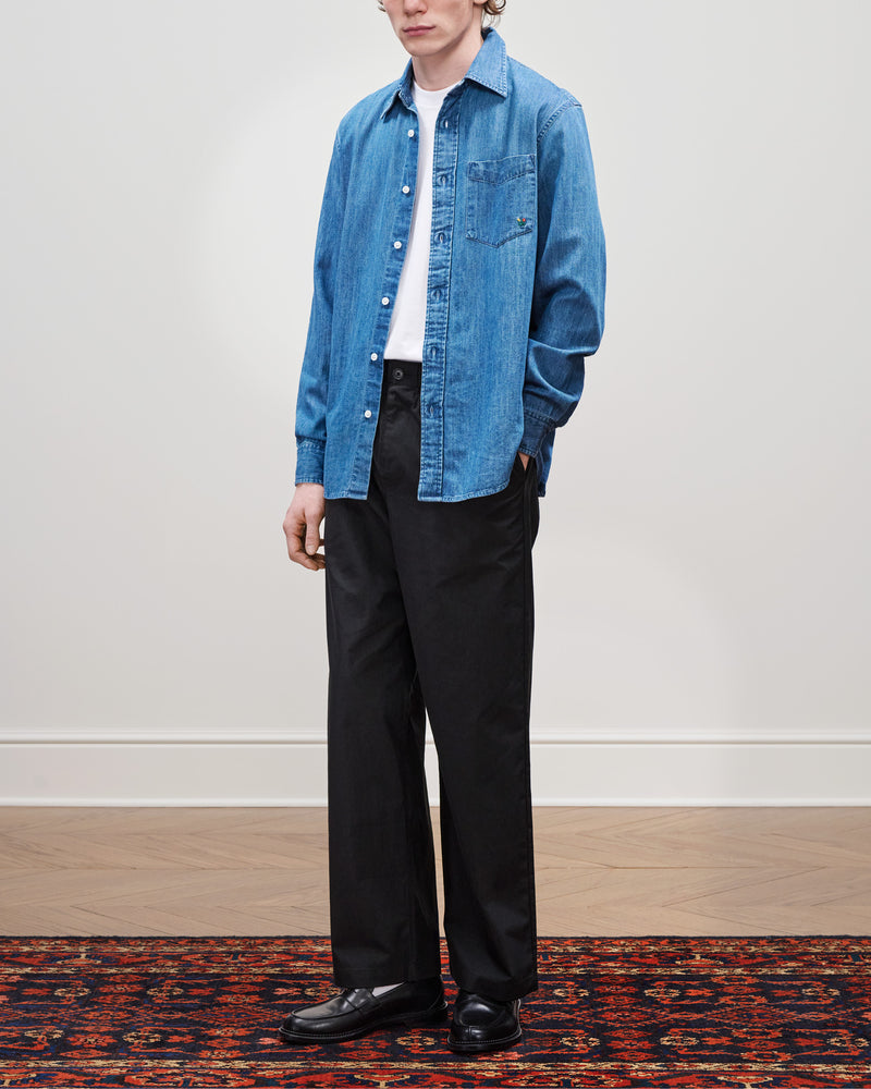 A person in a Crest Chambray Shirt, white tee, black trousers, and black shoes stands on a patterned rug.