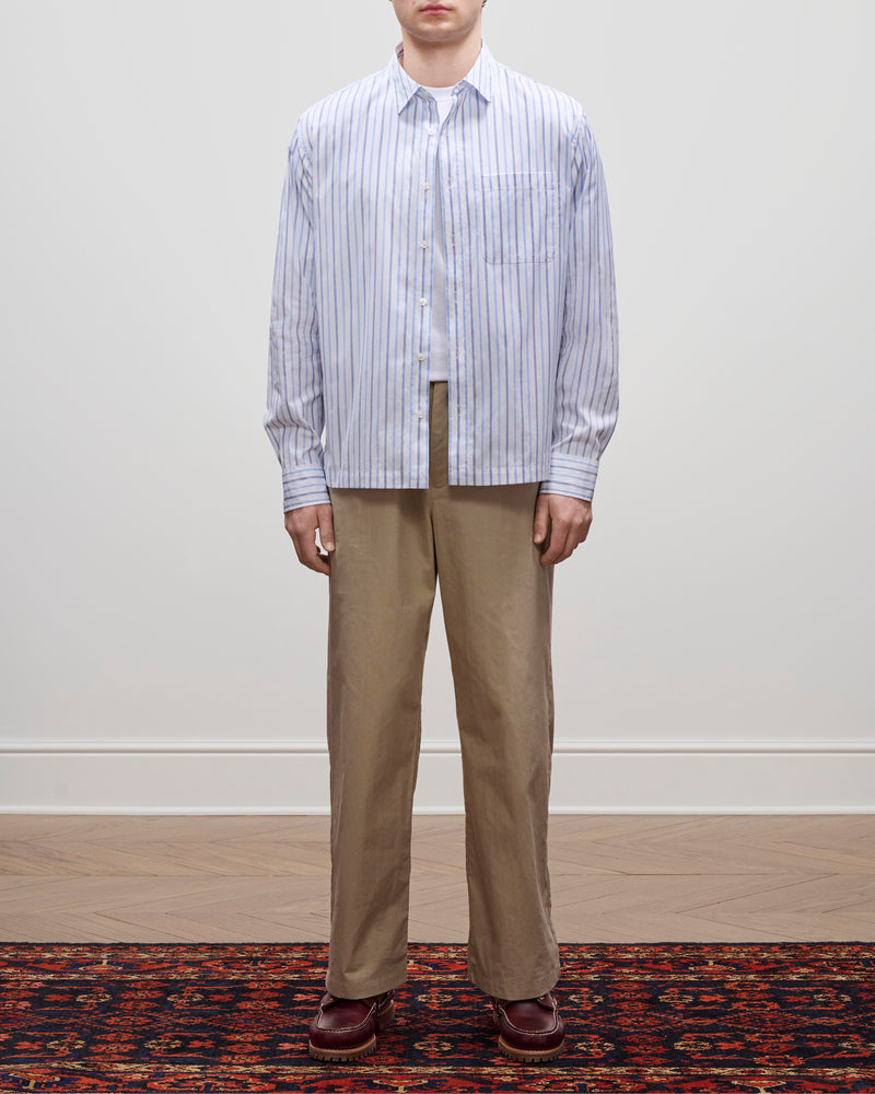 A person in Relaxed Cotton Trouser, a blue-striped shirt, and burgundy shoes stands on a patterned rug by a plain wall.