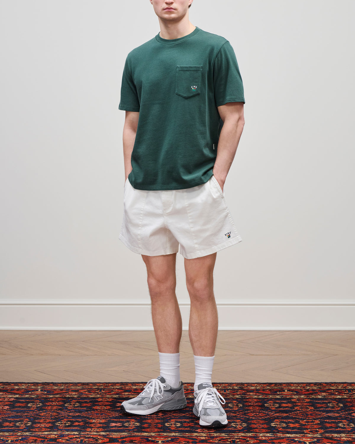 A person wears a Crest Pocket Tee, white shorts, gray sneakers, and white socks on a patterned rug.
