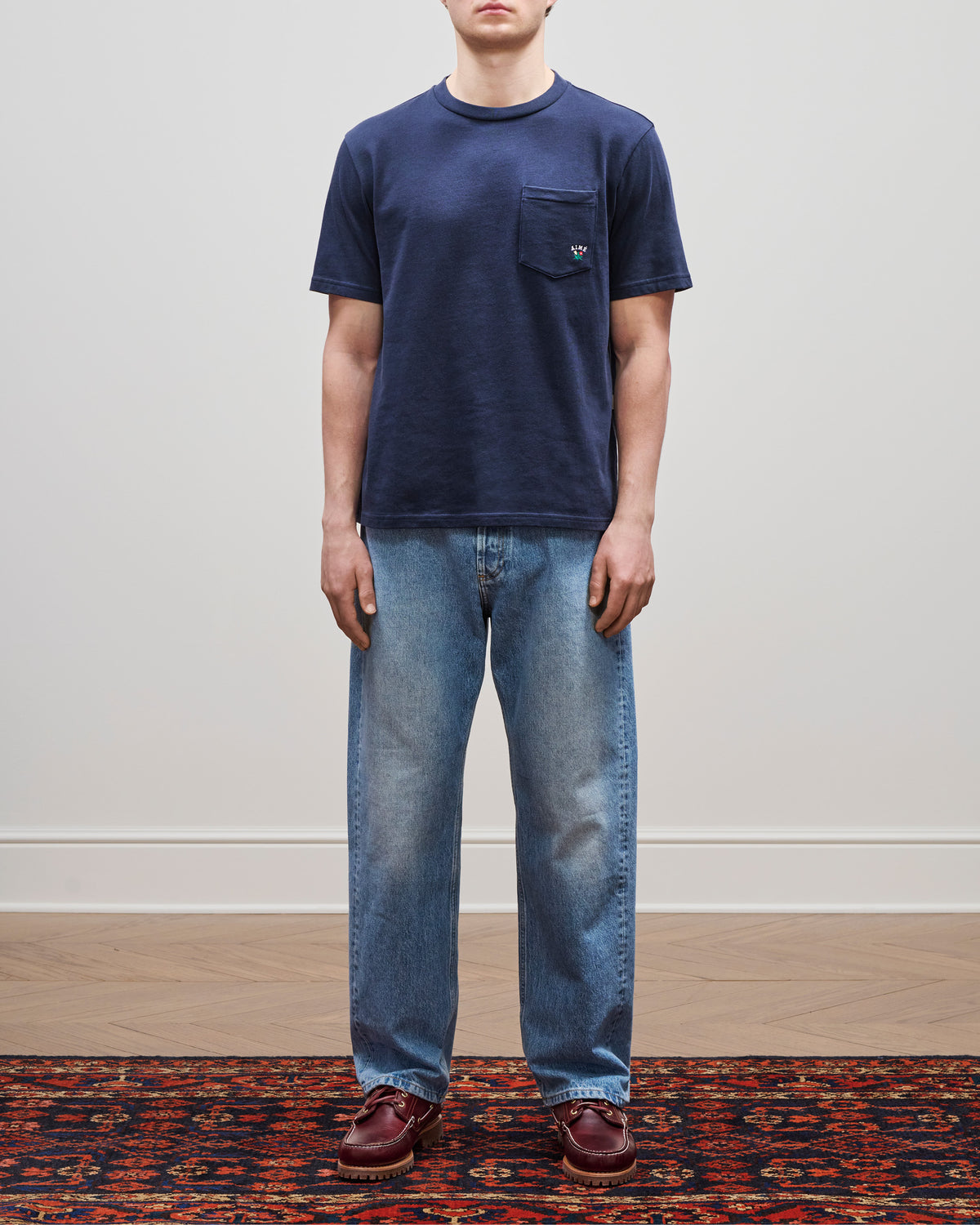 A person in a Crest Pocket Tee stands on a patterned rug against a plain wall.