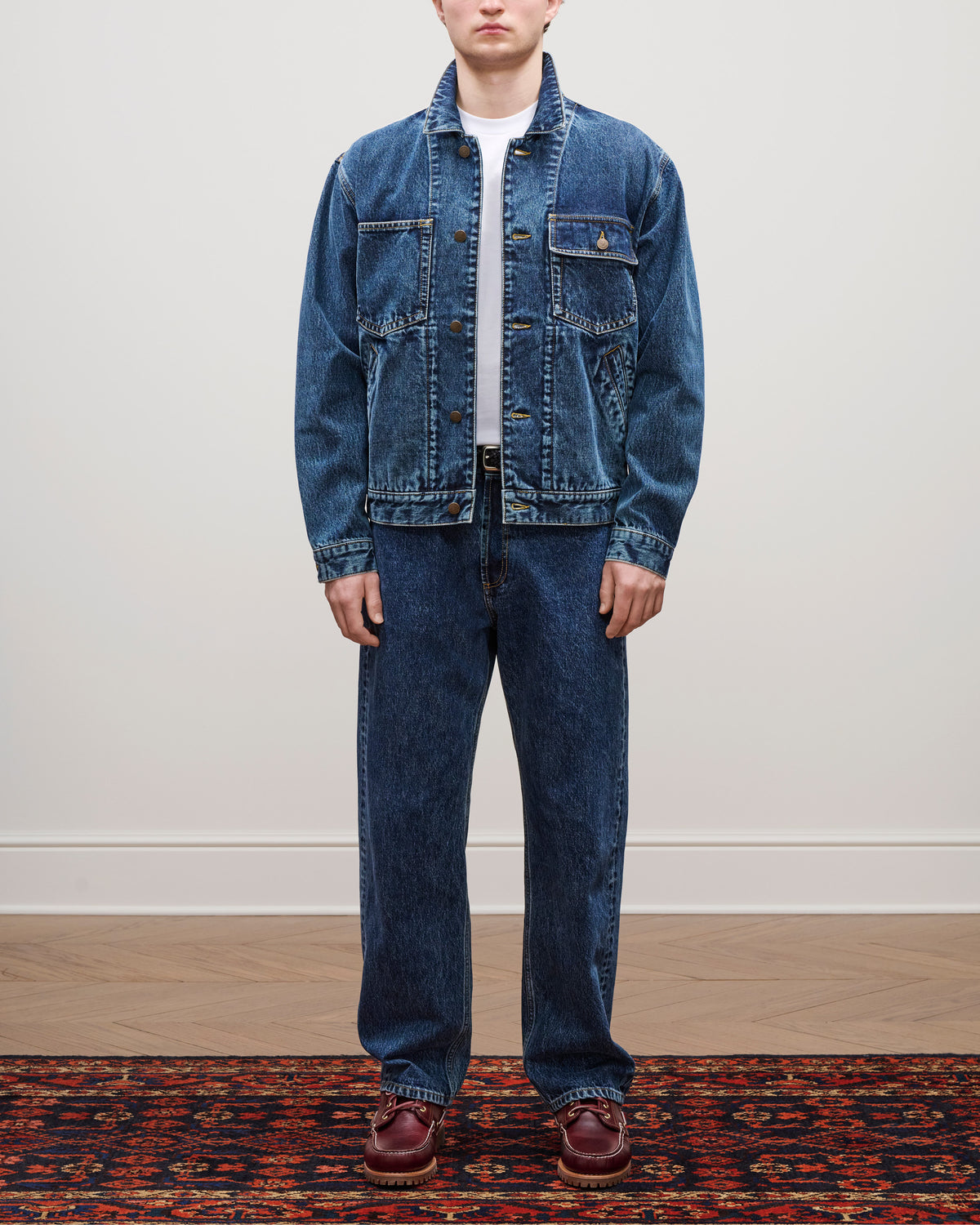 A person in a Denim Trucker Jacket, matching jeans, white shirt, and brown shoes stands on a patterned rug.