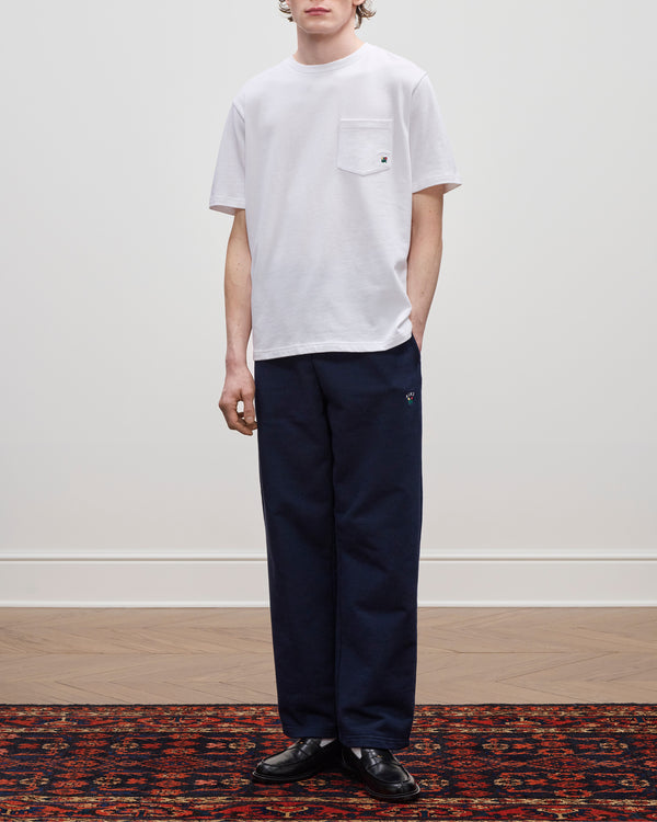 Someone wearing a Crest Pocket Tee in white and navy pants stands on a patterned rug against a plain wall.