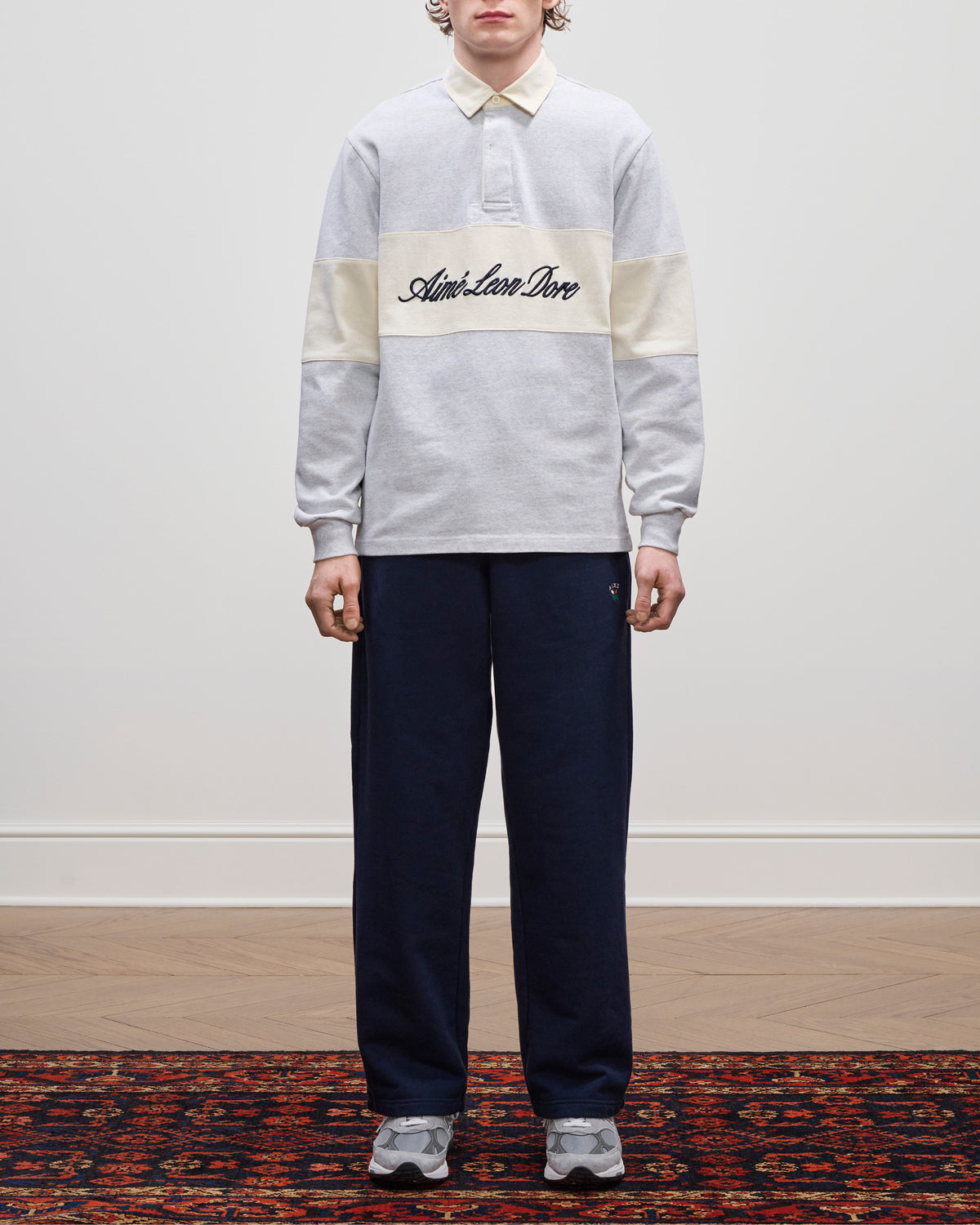 Person standing on a patterned rug, wearing the Script Paneled Rugby, navy pants, and gray sneakers.